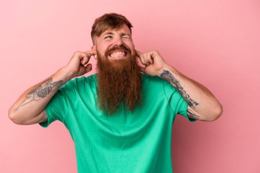 Uzun sakallı, beyaz, genç bir adam. Pembe arka planda izole edilmiş. Kulaklarını elleriyle kapatıyor..