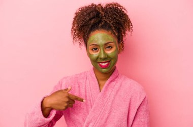 Bornoz ve yüz maskesi takan genç Afro-Amerikalı bir kadın pembe arka planda izole edilmiş ve elle bir tişört fotokopi alanını işaret ediyor, gururlu ve kendinden emin.