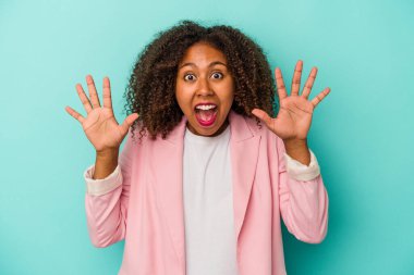 Mavi arka planda yalıtılmış kıvırcık saçlı genç Afrikalı Amerikalı kadın hoş bir sürpriz yaşıyor, heyecanlı ve el kaldırıyor..