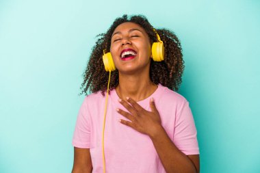 Mavi arka planda izole edilmiş müzik dinleyen genç Afrikalı Amerikalı kadın elini göğsüne koyup kahkahalar atıyor..