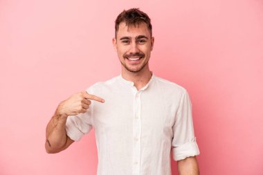 Pembe arka planda izole edilmiş genç beyaz bir adam gömlek fotokopi alanını elle gösteriyor, gururlu ve kendinden emin.