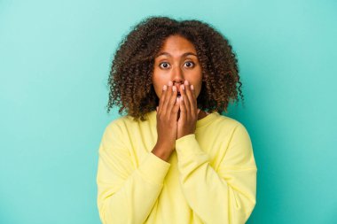 Mavi arka planda yalıtılmış kıvırcık saçlı, genç Afrikalı Amerikalı kadın elleriyle ağzını kapatırken şok oldu..