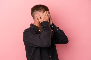 Uzun sakallı, beyaz, genç bir adam pembe arka planda izole edilmiş. Gözleri elleriyle kapamaktan korkuyor..