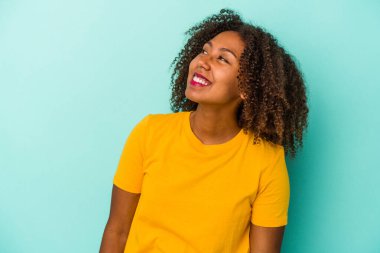 Mavi arka planda izole edilmiş kıvırcık saçlı, genç Afrikalı Amerikalı kadın. Rahatlamış ve mutlu gülüyor, boynunda dişlerini gösteriyor..