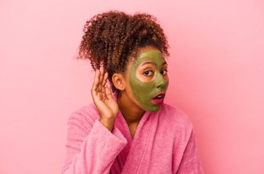 Pembe arka planda, bornoz ve yüz maskesi takan genç Afrikalı Amerikalı kadın bir dedikoduyu dinlemeye çalışıyor..