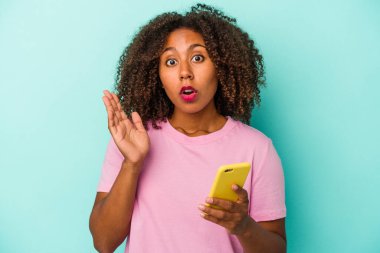 Elinde cep telefonu olan genç bir Afrikalı Amerikalı kadın mavi arka planda izole edilmiş. Şaşırmış ve şok olmuş..