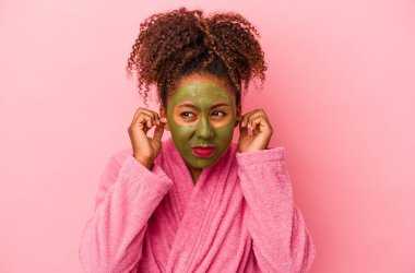 Bornoz ve yüz maskesi takan Afro-Amerikalı genç bir kadın. Pembe arka planda izole edilmiş. Elleriyle kulaklarını kapatıyor..