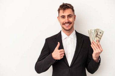 Beyaz arka planda izole edilmiş banknotları tutan Kafkasyalı genç iş adamı gülümsüyor ve başparmağını kaldırıyor.