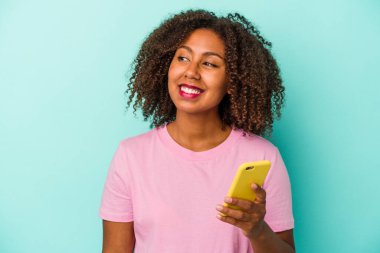 Mavi arka planda cep telefonu tutan Afrikalı genç bir Amerikalı kadın gülümsüyor, neşeli ve keyifli görünüyor..