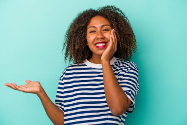 Mavi arka planda izole edilmiş kıvırcık saçlı Afro-Amerikalı genç bir kadın avuç içinde fotokopi alanı tutuyor, el üstünde tutuyor. Şaşırmış ve mutlu..