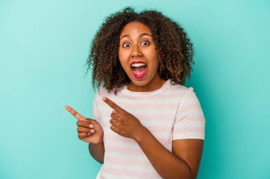 Mavi arka planda izole edilmiş kıvırcık saçlı, genç Afrikalı Amerikalı kadın işaret parmaklarıyla işaret ederek heyecanlanıyor..