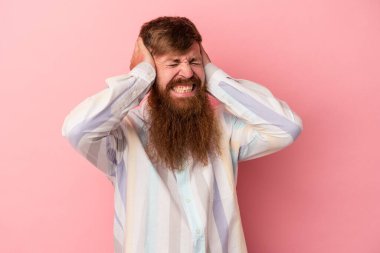 Uzun sakallı, beyaz, genç bir adam. Pembe arka planda izole edilmiş. Kulaklarını elleriyle kapatıyor..