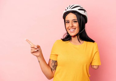 Tek kollu beyaz bir kadın pembe arka planda izole edilmiş bisiklet kaskı takıyor gülümsüyor ve boş alanda bir şey gösteriyor..
