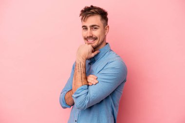 Pembe arka planda izole edilmiş genç beyaz adam gülümsüyor ve kendinden emin, elleriyle çenesine dokunuyor..