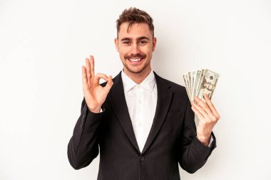 Kafkasyalı genç bir iş adamı elinde banknotlarla beyaz arka planda izole edilmiş neşeli ve kendinden emin bir şekilde iyi niyet gösteriyor..
