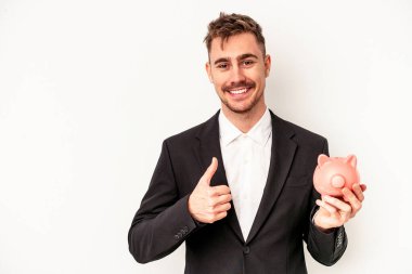 Beyaz arka planda izole edilmiş domuz kumbarası tutan Kafkasyalı genç işadamı gülümsüyor ve başparmağını kaldırıyor.