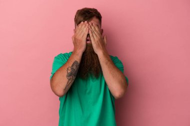 Uzun sakallı, beyaz, genç bir adam pembe arka planda izole edilmiş. Gözleri elleriyle kapamaktan korkuyor..