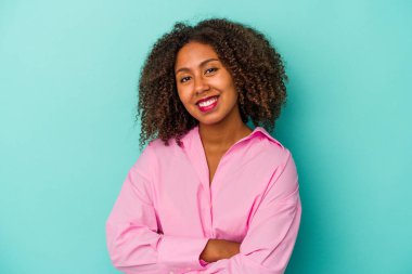 Mavi arka planda izole edilmiş kıvırcık saçlı genç Afro-Amerikalı kadın, kararlılıkla kollarını kavuşturuyor..