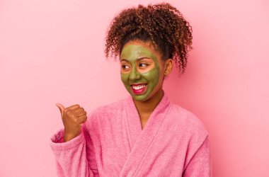 Bornoz ve yüz maskesi takan Afro-Amerikalı genç bir kadın pembe arka plan noktalarına izole edilmiş, başparmak uzakta, gülerek ve kaygısızca.