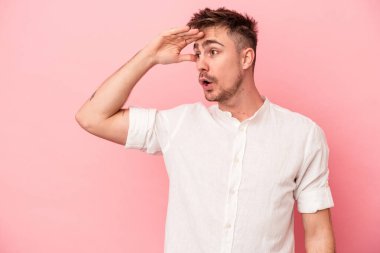 Pembe arka planda izole edilmiş genç beyaz adam uzaklara bakıyor ve elini alnına koyuyor..