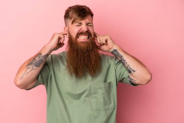 Kızıl saçlı, uzun sakallı genç adam pembe arka planda izole edilmiş parmaklarıyla kulakları örtüyor, gürültülü bir ortam yüzünden stresli ve çaresiz.