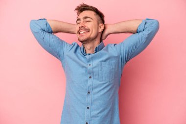 Pembe arka planda izole edilmiş genç beyaz bir adam kendini güvende hissediyor, elleri başının arkasında..