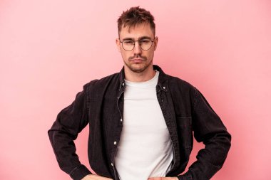 Pembe arka planda izole edilmiş genç beyaz adam hoşnutsuzca kaşlarını çatmış, kollarını kavuşturmuş..