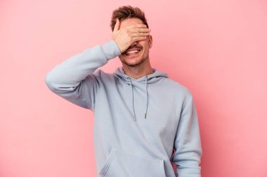 Pembe arka planda soyutlanmış genç beyaz adam elleriyle gözlerini kapatıyor, geniş bir gülümsemeyle bir sürpriz bekliyor..