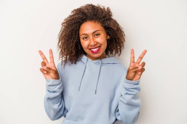 Beyaz arka planda yalıtılmış kıvırcık saçlı genç Afro-Amerikalı kadın zafer işareti gösteriyor ve gülümsüyor..