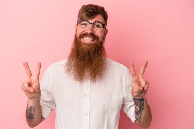 Pembe arka planda uzun sakallı beyaz bir adam zafer işareti gösteriyor ve gülümsüyor..