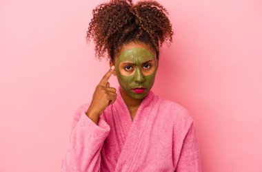 Bornoz ve yüz maskesi takan genç Afro-Amerikalı kadın pembe arka planda izole edilmiş, parmağıyla şakağı işaret ediyor, düşünüyor, bir göreve odaklanıyor..