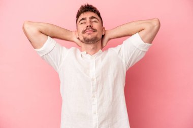 Pembe arka planda izole edilmiş genç beyaz bir adam kendini güvende hissediyor, elleri başının arkasında..