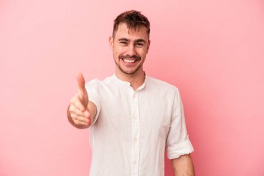 Pembe arka planda izole edilmiş genç beyaz adam selamlama jesti olarak kameraya elini uzatıyor..