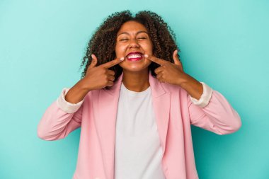 Kıvırcık saçlı, mavi arka planda izole edilmiş genç Afrikalı Amerikalı kadın gülümsüyor, parmaklarını ağıza doğrultuyor..