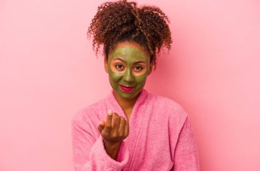 Genç Afro-Amerikalı kadın bornoz ve yüz maskesi takıyor pembe arka planda izole edilmiş seni işaret ediyor sanki davet ediyormuş gibi..