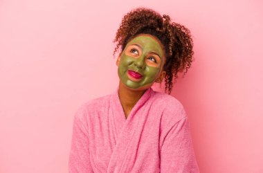 Bornoz ve yüz maskesi takan Afro-Amerikalı genç bir kadın pembe arka planda izole edilmiş hedeflere ve amaçlara ulaşma hayali kuruyor.