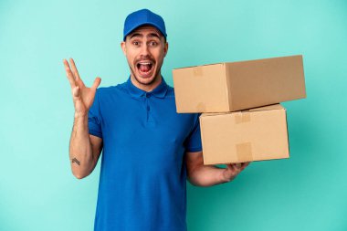 Genç teslimatçı beyaz adam mavi arka planda izole edilmiş hoş bir sürpriz alıyor, heyecanlı ve el kaldırıyor..