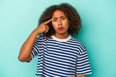 Mavi arka planda izole edilmiş kıvırcık saçlı, genç Afrikalı Amerikalı kadın şakağı parmağıyla işaret ediyor, düşünüyor, bir göreve odaklanıyor..