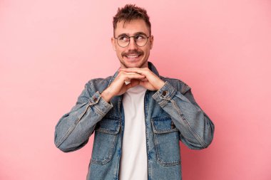 Pembe arka planda izole edilmiş genç beyaz adam ellerini çenesinin altında tutuyor, mutlu bir şekilde yana bakıyor..