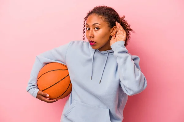 Young african american man playing basketball isolated on pink background trying to listening a gossip.