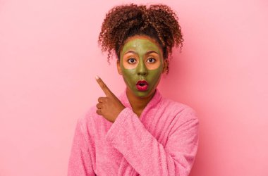 Üzerinde bornoz ve yüz maskesi olan genç bir Afro-Amerikalı kadın pembe arka plana izole edilmiş.