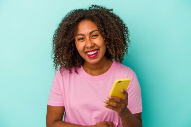 Genç Afrikalı Amerikalı bir kadın elinde cep telefonu tutarken mavi arka planda izole bir şekilde gülüyor ve eğleniyor..