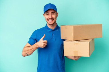 Genç teslimatçı beyaz adam mavi arka planda izole edilmiş gülümsüyor ve başparmağını kaldırıyor.