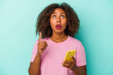 Genç Afro-Amerikalı bir kadın cep telefonunu mavi arka planda izole edip ağzı açık bir şekilde işaret ediyor..