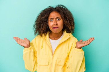 Mavi arka planda yalıtılmış kıvırcık saçlı, genç Afrikalı Amerikalı kadın. Kopya alanını tutmak için omuz silkme konusunda kafası karışık ve kuşkulu..