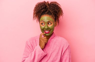Bornoz ve yüz maskesi takan genç Afro-Amerikalı kadın pembe arka planda izole edilmiş, rahat bir şekilde fotokopi odasına bakan bir şeyi düşünüyor..