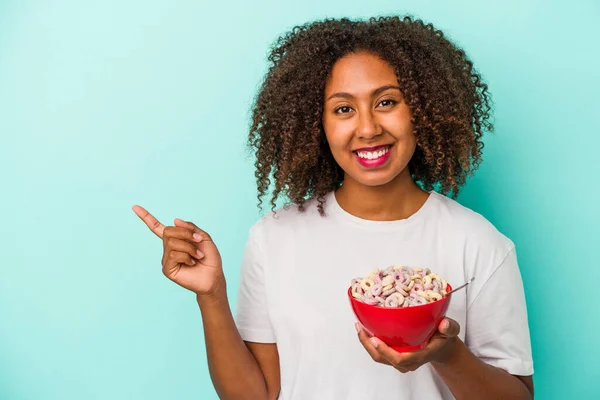 年轻的非洲裔美国女人拿着一碗谷物 蓝色背景隔离 微笑着指着旁边 在空白的空间里展示着一些东西 — 图库照片