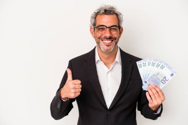 Orta yaşlı iş adamı mavi arka planda izole edilmiş banknotları tutuyor gülümsüyor ve başparmağını kaldırıyor.