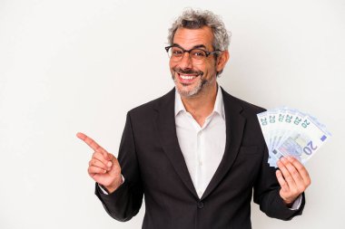 Orta yaşlı bir iş adamı mavi arka planda izole edilmiş banknotlar tutuyor gülümsüyor ve boş alanda bir şeyler gösteriyor..