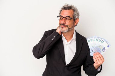 Elinde banknotlar tutan orta yaşlı bir iş adamı mavi arka planda, kafasının arkasına dokunup düşünüyor ve bir seçim yapıyor..